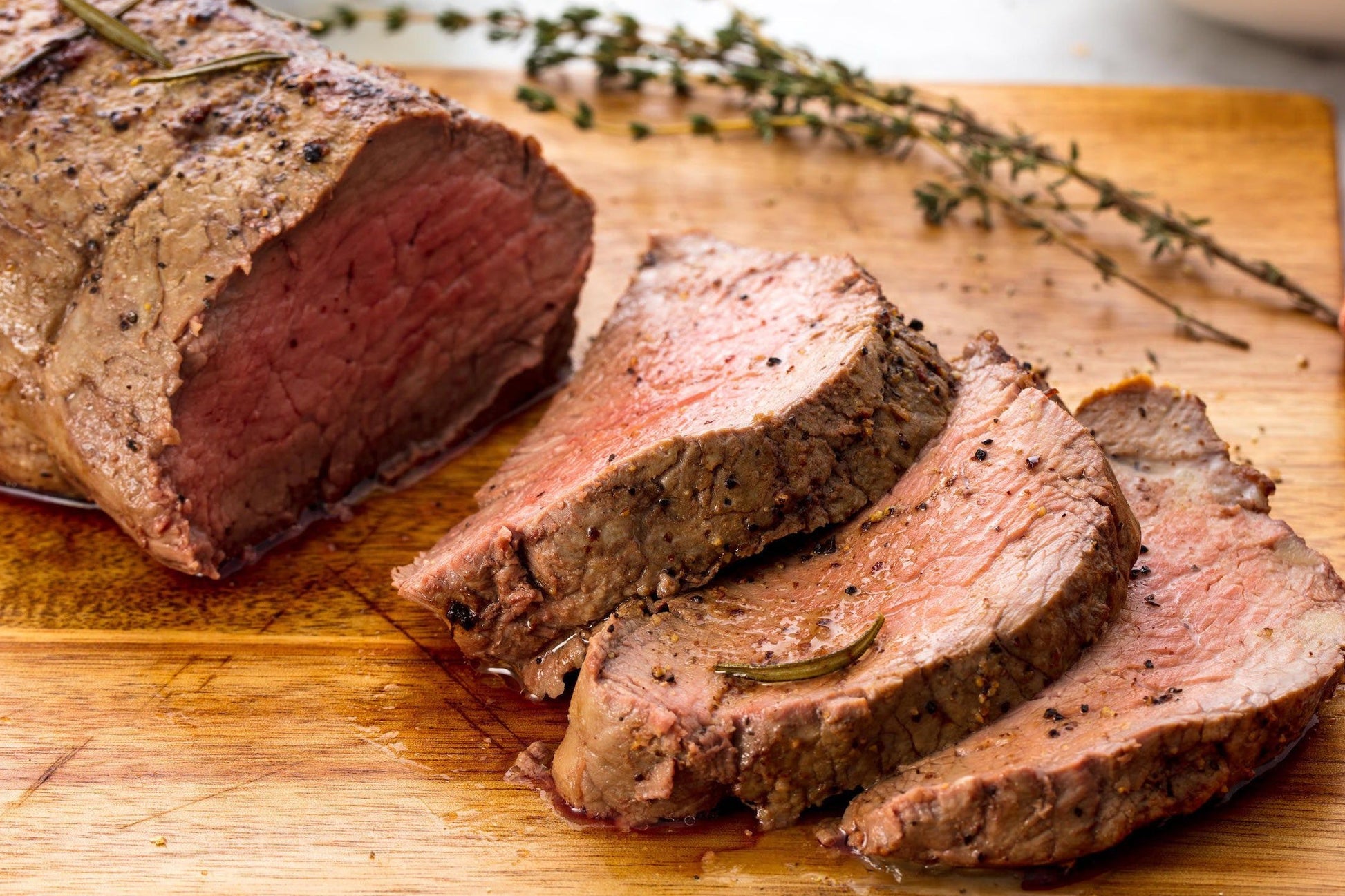A cooked beef tenderloin steak on a wooden cutting board with a knife, surrounded by sliced steak and herbs.